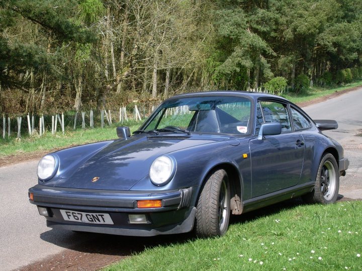 Pictures of your classic Porsches, past, present and future - Page 27 - Porsche Classics - PistonHeads - The image displays a blue sports car parked on what seems to be a well-trodden, grassy area at the side of a road. The car is facing the left side of the image, giving a clear view of its side and rear profile. The vehicle's bodywork appears sleek and well-maintained, with a prominent air intake above the rear wheel arch. There is a new-style Porsche logo on the front of the car, along with a license plate reading "FE7 GNT" that is partially visible. The setting suggests a rural or suburban environment, with a fence and trees lined along the road behind the car. The overall appearance of the car and its surroundings conveys a sense of tranquility and enjoyment of the open road.