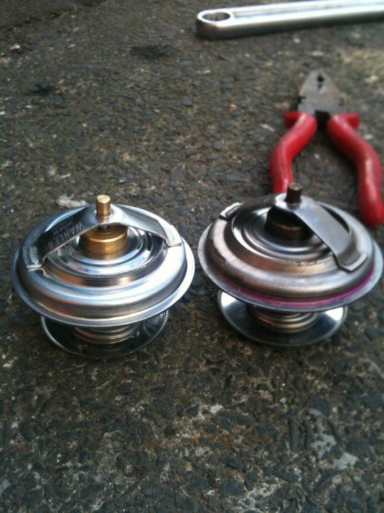 Today Pistonheads Cleaned - The image showcases a pair of specialized barbells placed on an asphalt surface. The barbell weights bear a metallic sheen with machined edges, and are separated by a fabric Central Bar. A clamp with red, curved handles, partially open, is situated next to the barbell weights, suggesting a recent repositioning or serving as a tool for movement or adjustment. The presence of the clamp in the frame indicates this might be a preparation or finishing area for weightlifting equipment.