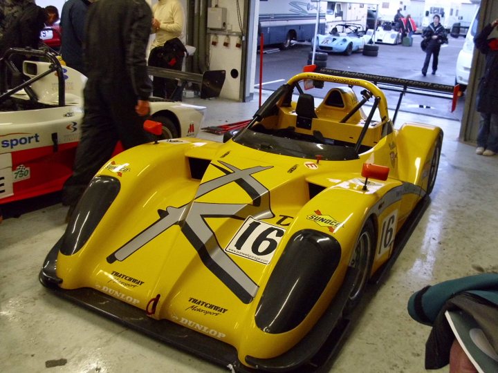 Association Day Picturs Stroke Pistonheads - The image showcases a yellow racing cockpit viewed from behind, prominently displaying the number 16. Adjacent to the cockpit, a man in a dark suit walks, showing his back as he moves away. In the background, several other race cars are visible, indicating a garage or paddock setting. Exiting the scene, a person in a dark coat can be seen, while the setting is open and spacious, typical of a racetrack area. The environment suggests a racing or sporting event.