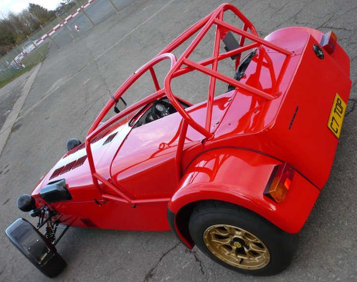 Pistonheads - The image showcases a small, red, single-occupant sports car with a prominent rear-wing spoiler. It's situated on a pavement parking lot, near a fence, appearing to be either arriving or departing from the static image. The car is equipped with gold rims, which contrast richly with the red body.