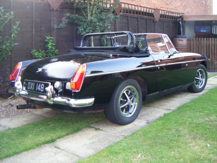 Rubber to Chrome Bumper Conversion - Page 1 - MG - PistonHeads - The image features a classic dark green, possibly black, convertible parked on a brick driveway. Behind the car, a wooden fence can be seen, implying a residential setting. The vehicle has a convertible top up and is equipped with wire wheels and red taillights. To the left of the photo, potted plants are visible, adding a touch of greenery to the scene. The overall scene conveys a sense of style and history, harkening back to the classic convertible era.