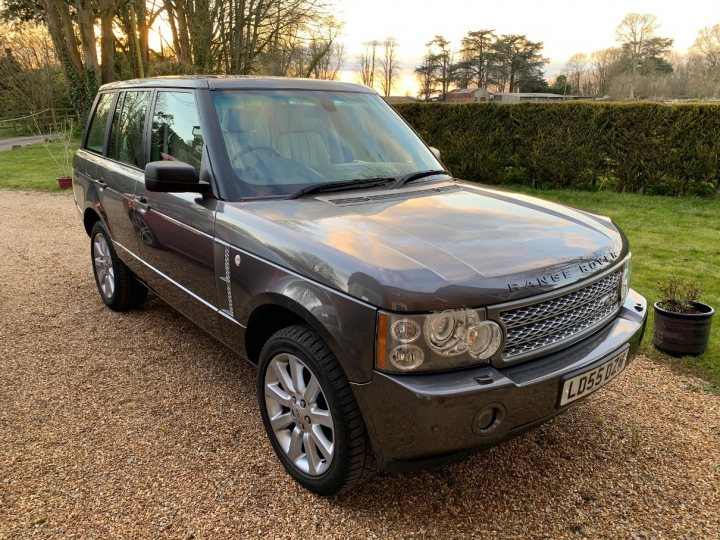 A truck parked on the side of a road - Pistonheads - This image features a parked Land Rover vehicle. The car is silver and appears to be in good condition, with no visible damage or scratches. It has the standard design of a Range Rover, including the characteristic boxy shape and elevated ride height. In front of the car, there are what seem to be two potted plants. There's also a person present in the background, but only a small part of their body is visible. The setting appears to be outdoors during daylight hours, as suggested by the natural lighting and the presence of trees and grass in the vicinity.