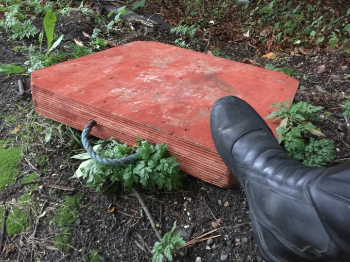 Odd stuff you have hit or avoided in the road. - Page 1 - Biker Banter - PistonHeads - The image shows a rusted red piece of metal lying on the ground in an outdoor setting. The background features a mix of natural elements such as trees and plants, suggesting this scene is in a park or similar green space. To the left, there's a boot touching the edge of the metal piece, indicating someone is examining it. There are no people fully visible in the image, and only a portion of a foot is seen interacting with the metal object. The overall atmosphere suggests an exploration or discovery moment.