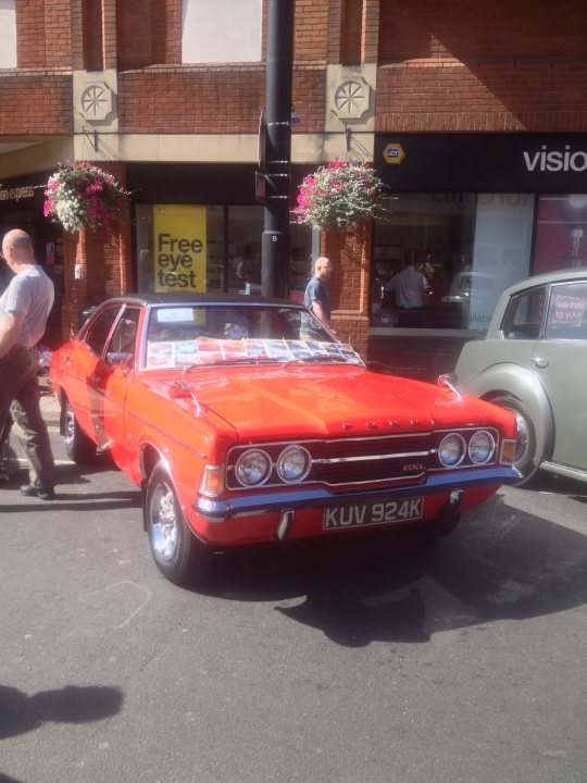 A red car parked in front of a building - Pistonheads - The image features a vibrant red car parked prominently on the street. It's a classic model with distinctive round headlights, epitomizing a bygone era. The car is parked outside of a store, adding a sense of everyday life to the scene. In the background, there's a gray car parked alongside other vehicles, and pedestrians are visible, going about their day. Scattered around the area are a couple of potted plants, adding a touch of greenery to the urban landscape.