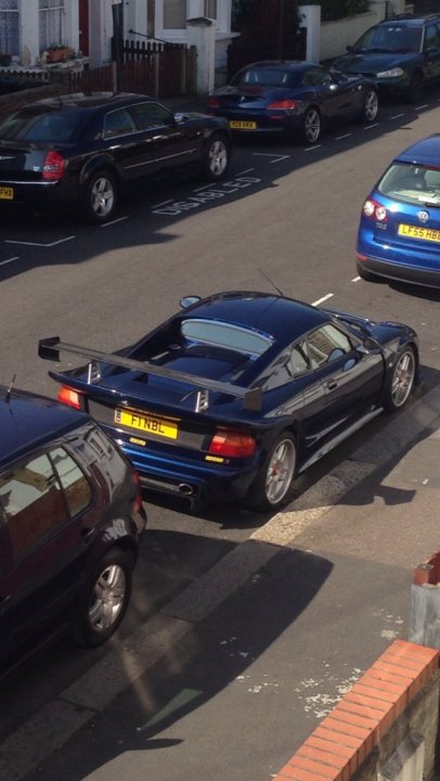 Pistonheads - The image depicts a vibrant street scene with a primary focus on several luxury cars parked along the side of the road. These cars, which include models from various automakers, are parked in front of a parking bay marker. In the foreground of the image, there is a distinctive black sports car, possibly a Lamborghini, parked adjacent to a standard hatchback car. In the background, various other vehicles, including a larger blue van, can be seen adding to the bustling urban atmosphere.