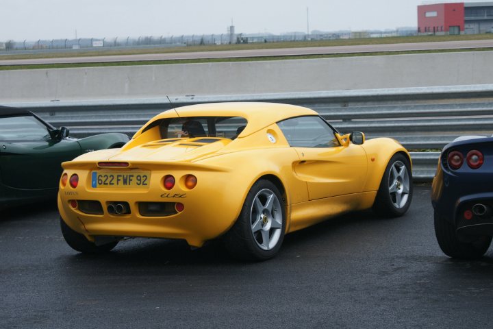 French Photos - Page 1 - Elise/Exige/Europa/340R - PistonHeads - In this image, the main focus is a yellow sports car that is a 2004 Lotus Elise, evident from its distinctive red tail lights. It's parked in a parking lot that also features a few other cars. The play of light and shadow highlights the car's features, including its sleek design, round headlights, and the time "10:07" displayed on the top edge of the image. The setting seems to be on a cloudy day with an industrial background visible in the top right.