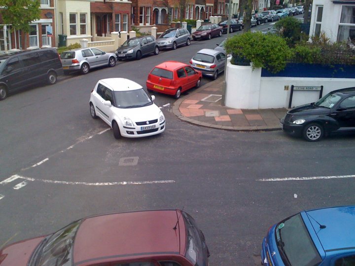 Pistonheads - This is an aerial view of a residential street showing a variety of parked cars. The street appears to have two parking lanes on each side, with parked cars facing different directions. The entrance to a residential driveway is also clearly visible. The cars are in different colors, including black, red, blue, and white. The number of cars suggests a busy neighborhood.