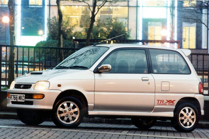 Calling all unusual hot hatch owners - Page 6 - General Gassing - PistonHeads - The image shows a small white car parked on a street at night. The car is a hatchback with a visible spare tire on the rear door. There are stylish alloy wheels and a pair of antennas on top. Behind the car, there is a well-lit building, and the lighting suggests it's evening or nighttime. The car's lights are on, which contrasts with the darker surroundings. The scene is quite naturalistic, capturing a moment of stillness on the road at night.