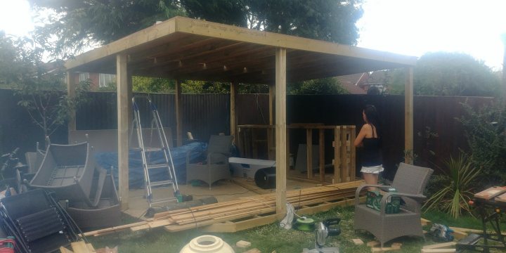 My Garden Bar - Page 1 - Homes, Gardens and DIY - PistonHeads - The image captures a scene in a backyard where construction work is underway. A large patio area, featuring wooden planks and metal framing, dominates the view. Workers can be seen amidst the materials scattered on the ground, suggesting an ongoing project. There's a sense of activity as various tools and equipment are visible. In the background, a partially constructed structure looms, hinting at the scale and complexity of the work.