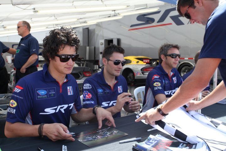 New GTS-R race debut - Page 1 - Vipers - PistonHeads - The image shows a group of men at an autograph signing event. They are seated at a table, with one man standing and leaning over another. The table is covered with marketing materials and sports merchandise. All the men are wearing matching blue jerseys, suggesting they are part of the same team. The most prominent team's name on their jerseys is "SRRT." The setting appears to be outdoors, under a tent-like structure, as indicated by the presence of "SRRT" on the tent's banner.