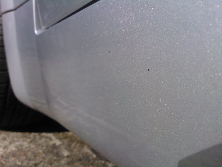 Pistonheads - The image is a close-up view of a car's body panel, specifically the lower corner of its rear side door. The angle is from the bottom and slightly angled towards the front of the vehicle, focusing on a particular spot where the paint appears slightly brushed or damaged. There is also a black object, possibly a fleck, near a seam in the metal, which could be interpreted as a blemish or scratch. The texture of the car's finish indicates that it might not have a high-gloss protective layer, but rather a standard clearcoat. The background is blurred, emphasizing the car's paint and the area of interest.