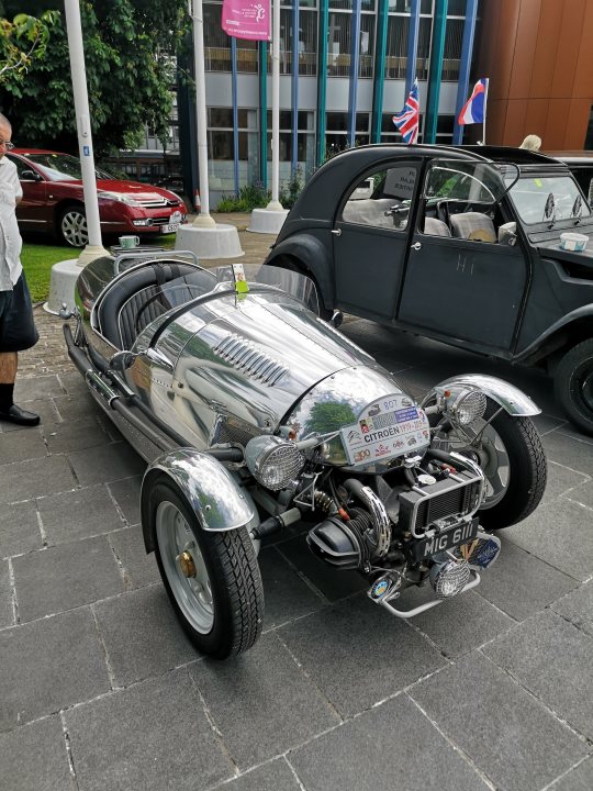 A motorcycle parked on the side of a road - The image showcases a classic car, which is the focal point. It has an old-fashioned design, reminiscent of classic sports cars from a past era. The car is parked on the street next to a building, suggesting it might be part of a display or event. In the background, there are several other cars and people present, adding context and activity to the scene. There's also a flag visible in the background, possibly indicating the location or an ongoing event.