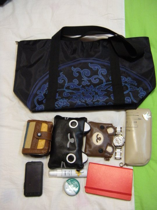 This image captures a collection of personal items neatly arranged on a surface. The items include a black handbag with a distinctive blue design, a brown leather wallet, a red phone case, a watch, a pair of eyeglasses, two pens, a cosmetic case, and three scent sticks. The objects are spread out to showcase each item, suggesting they might be part of a travel or everyday carry-all set. The layout is organized, with the wallet and glasses placed next to each other while the cosmetic case and scent sticks are situated close to the handbag. The setting could be a bed with a glimpse of a green blanket or sheet in the background.