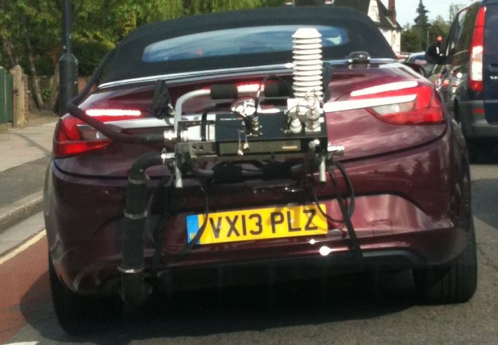 New Vauxhall? - Page 1 - VX - PistonHeads - The image presents a close-up view of the back of a maroon-colored car on a road. The car is equipped with a trailer combined with a car carrier device attached to its rear. The trailer is equipped with a spiral flexible hose, which might be a part of a performance enhancing system. The license plate on the car reads "VXI 3 PZ." The setting appears to be a street with a curb, cars are visible in the background.