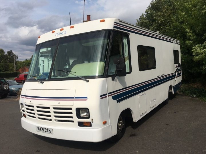 A class Vs coachbuilt motorhome  - Page 1 - Tents, Caravans & Motorhomes - PistonHeads - The image shows a white RV parked in an open space. It features the text "Trophy" on the side and has a blue stripe running across its length. The RV is positioned facing the viewer, revealing its interior through the large windows. In the background, there's another smaller vehicle to the left of the RV, partially obscured by it. The sky is visible in the top part of the image, suggesting an outdoor setting.