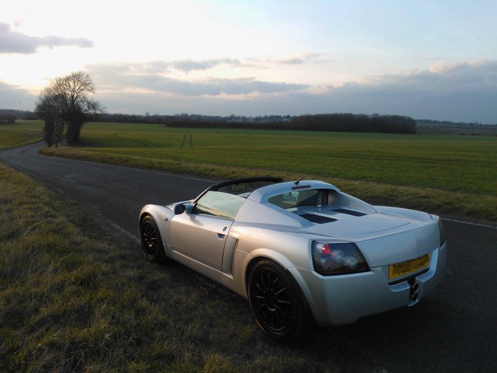 Pistonheads - The image showcases a sleek, silver sports car parked on the side of a road. The road curves gently to the right, hinting at an open landscape beyond. On the left, a lone tree stands, its leaves a darker shade of green. Above, the sky is a vast expanse of clouds, suggesting that it might be an overcast day. The setting appears serene, with the car as the main point of interest.