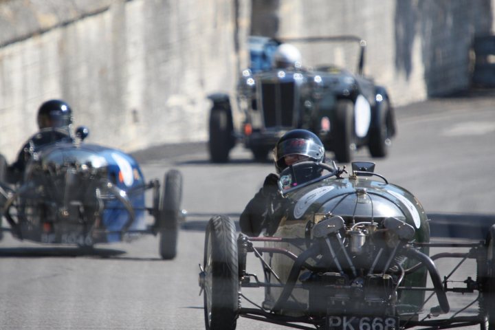 Making Pistonheads Wheelers - The image captures a dynamic scene of vintage race cars on a paved straightaway. Two cars are visible, one leading and bearing the number "PK 668," the other slightly behind and slightly left. Both drivers are wearing helmets, and the track is bordered by a contrasting out-of-focus photograph. The cars' unique designs and metallic surfaces gleaming in the light suggest the intensity of the race.