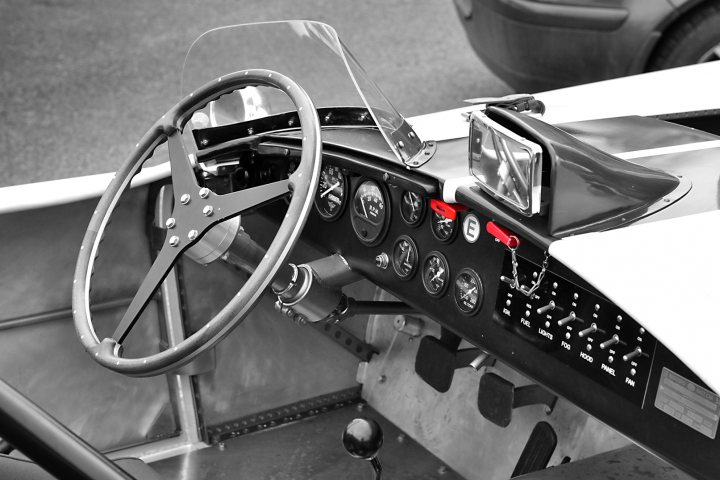 Robert Barrie Track Day 24th Feb Goodwood - photos - Page 1 - South Coast - PistonHeads - The image features a vintage racing car with a prominent steering wheel at the center of the frame. The steering wheel appears to be made of leather with silver-colored metal accents. Surrounding the steering wheel are multiple dials and gauges, indicative of the car's controls for speed, acceleration, and other essential measures. The background is blurred, but it seems to be an outdoor setting with a partially visible vintage vehicle, suggesting the photograph was taken in a collection or exhibition where classic cars are displayed. The overall impression is of a well-maintained, classic race car ready for the track, with the engine and dashboard in focus.