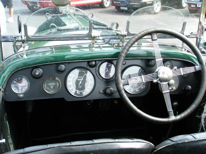 Breakfast Meeting, The Hare, Roxwell 4th June - Page 1 - Kent & Essex - PistonHeads - The image displays the interior of an old, classic car. The car has a distinctive black and white steering wheel, with numerous gauges and controls. The most prominent gauge is in the center of the steering wheel, with two other gauges positioned on the sides. The steering wheel also has a unique emblem in its center. The dashboard behind the steering wheel is adorned with a unique design, and the overall interior of the car suggests a blend of comfort and vintage charm, enhanced by the vintage aesthetic of the gauges and controls. The colors in the image are primarily black, white, and green, which is typical for classic cars.