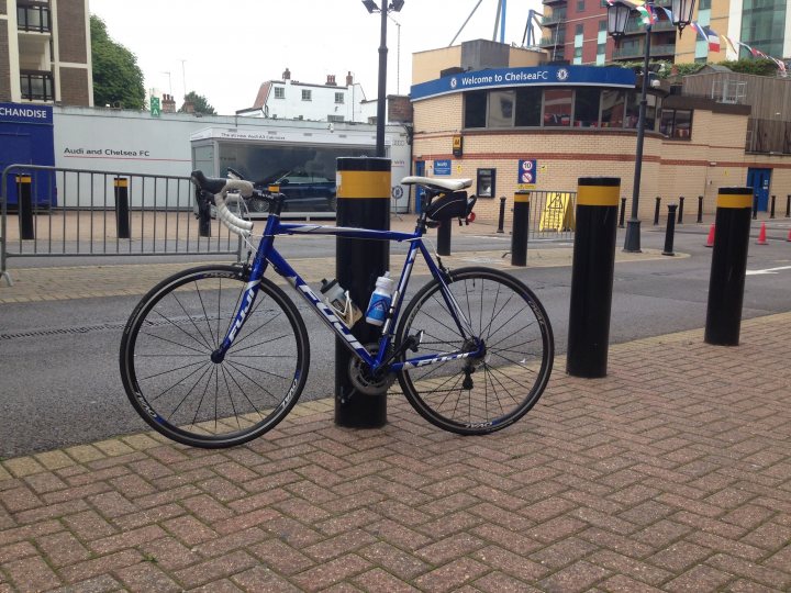 The "Photos From Today's Ride" thread... - Page 150 - Pedal Powered - PistonHeads - The image features a beautiful blue bicycle parked on a brick sidewalk. The bicycle is positioned next to a black metal post. In the background, there's a large, open space that seems to be a street or parking lot, with some yellow markings. The environment suggests an urban setting, possibly near a park or a public area. The bicycle appears to be stationary, possibly locked, indicating that the owner might have gone to take a break or run a quick errand nearby.