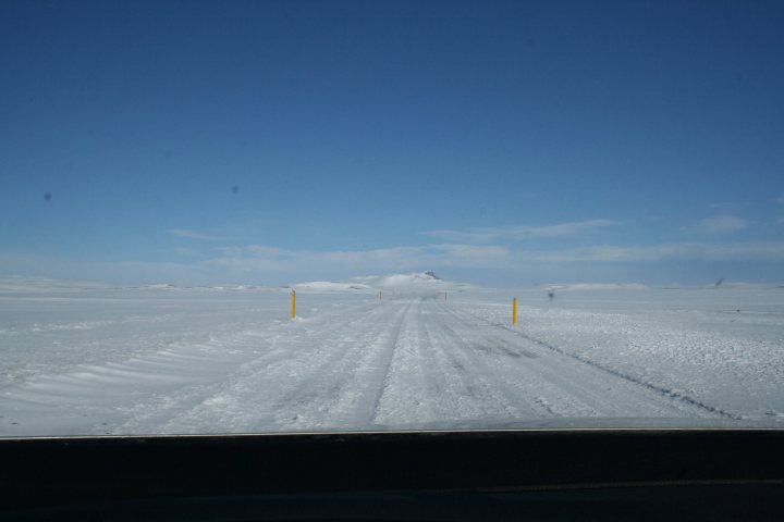 Iceland on a budget? - Page 1 - Holidays & Travel - PistonHeads - This image shows a snowy landscape in daylight. The perspective is from inside a vehicle, looking out towards the front windshield, which is visible in the foreground. The windshield is covered with snow, reflecting the bright, cloud-filled sky above. The flat terrain, rendered mostly in white due to the snow, stretches out to a distant horizon. In the middle distance, there are a few quaint yellow markers surrounding what looks like a small, unpaved track covered by snow; this track appears to be part of a remote roadway. There is no immediate sign of human activity or habitation, beyond the pole to which the markers are attached.