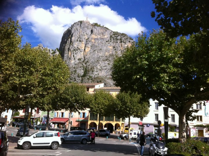 Driving France - Page 4 - Chimaera - PistonHeads - The image presents a small town scene with a commanding mountain in the backdrop. The town's streets are lined with parked cars, including a white compact car with a red light on top. A person is standing on the sidewalk, and there are trees lining the streets, providing a mix of shade and open views towards the mountain. The sky overhead is partly cloudy, suggesting a pleasant day. The overall atmosphere is calm and quaint, typical of a small rural town.