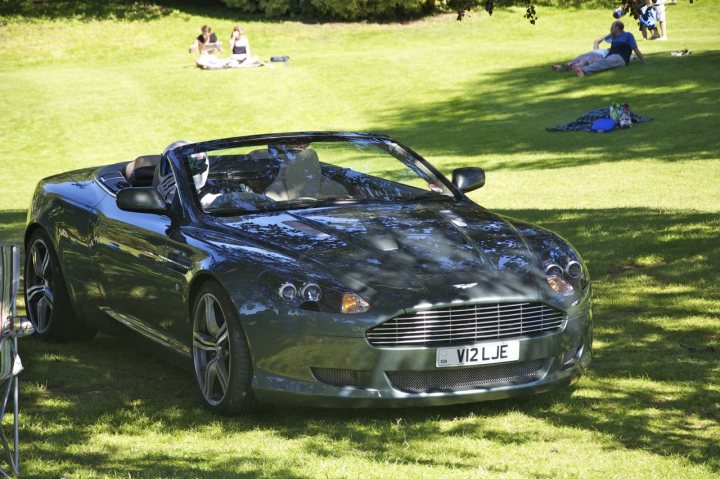 hever pics - Page 1 - Aston Martin - PistonHeads - In the image, there is a single, dark green sports car parked in a verdant field buzzing with people lounging and socializing. The car is gleaming under the sun, with its roof down and top up. The sunlit field extends to meet a line of trees in the distance, creating a tranquil scene.