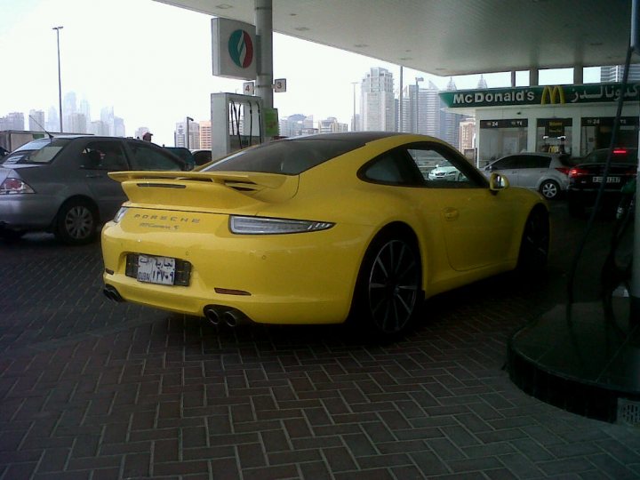 Middle East spotted thread - Page 50 - Middle East - PistonHeads - This image depicts a vibrant cityscape. The main focus is a bright yellow Porsche Cayman sports car, parked off to the side in front of a McDonald's. The car is sleek with a tilted spoiler, black wheels, and a black soft-top roof, exuding speed and style. It's a sunny day, and the sky is clear, reflecting off the chrome details of the car, making it shine brightly. The McDonald's in the background is illuminated against the sky, serving as a contrasting element in the scene.