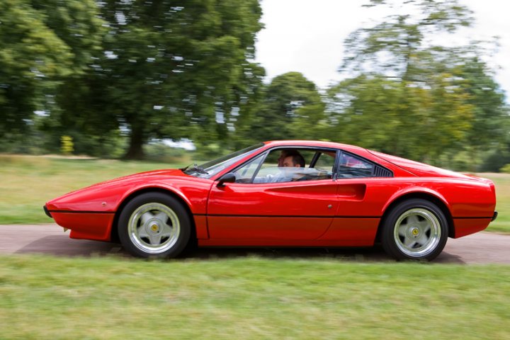 Pistonheads Manor Motors - The image showcases a vibrant red, open-top sports car speeding down a scenic road. The car is in motion, capturing the essence of speed and luxury. The car and its driver are the main focus of the image, contrasting with the serene backdrop of lush green trees and grass. The dash of color from the car against the natural hues of the greenery creates a striking visual contrast.