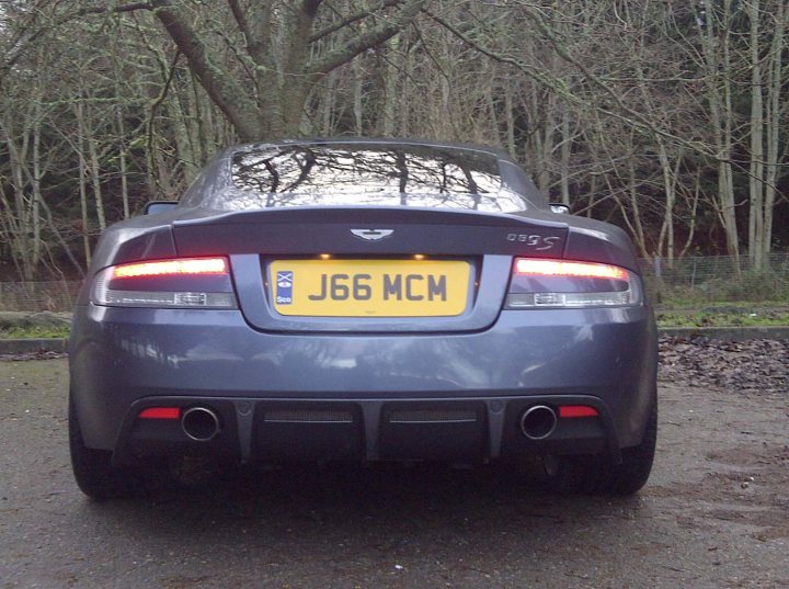 DBS wheels on DB9 - (more or less) - Page 1 - Aston Martin - PistonHeads - The image features a dark blue Jaguar car parked on a paved area surrounded by trees. A clear view of the car's rear end is provided, showing the brake lights, exhaust pipe, and the license plate which reads "J66 MCM". The car is shining under natural light, and a person can be seen in the distance, partially obscured by the foliage.