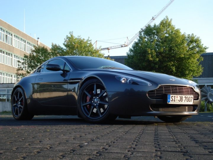 My first Aston - Page 1 - Aston Martin - PistonHeads - The image showcases a sleek and stylish black Aston Martin DB7, a luxury sports car renowned for its performance and design. The car is parked on a vibrant red brick ground, facing towards the right side of the frame. In the background, there's a building and a crane, suggesting an urban setting, possibly near an ongoing construction or development project. The sky above is clear, providing a beautiful contrast to the scene, highlighting the car's shiny, well-maintained appearance.