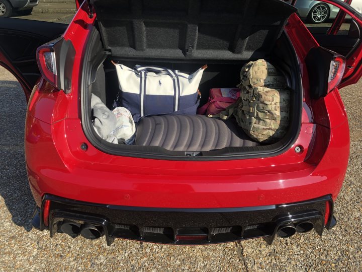 Type R - Page 1 - Readers' Cars - PistonHeads - The image shows a red sports car with its trunk open, revealing items inside. Among these items is a large white bag, possibly containing camping equipment, and a smaller black and white bag. A person's torso is visible in the back of the car, suggesting that someone might be in the process of loading or unloading the vehicle. The car is parked on an asphalt surface, and in the background, there's another car parked further away.