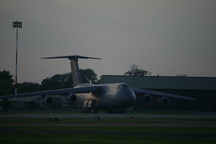 An airplane is parked on the runway at the airport - Pistonheads - The image shows an oversized gray airplane located on the tarmac of an airfield or airstrip. The airplane is massive, with multiple engines visible under its wing. Behind the airplane, there's a large, rectangular building with a flat roof, possibly an airport hangar or terminal. The lighting in the image seems to come from natural light, suggesting it could be either dawn or dusk.