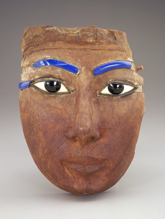 The image showcases a sculpture of a face, possibly of ancient Egyptian origin, made of a wood-like material. It prominently features blue painted eyebrows and eyes, with the eyes closed. The face is dark, highlighted with blue near the eyes, and has a distinctive nose and lips, giving it a distinct character. The sculpture appears to be an artistic representation rather than a realistic likeness.