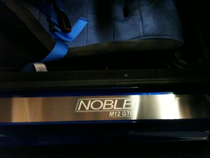 stainless sill plates - Page 1 - Noble - PistonHeads - The image shows a glimpse of the interior of a sports vehicle, featuring a dark blue car seat with a contrasting blue seat belt. The seat is positioned in such a way that it allows a view underneath the car seat, where a sleek, stainless steel rail or door hinge is visible, displaying "NOBLE M12 GT" in a silver oval logo. The focus is on the detail of the steel color accent and the name embossed on the vehicle's component. The ambiance suggests the photo may have been taken under low or dim lighting, as the interior of the vehicle is in shadow.