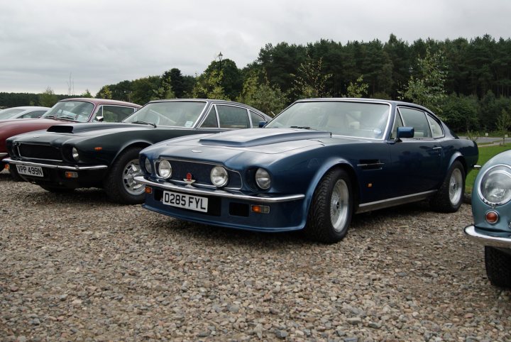 Astons & Ferraris at Glenfiddich distillery - Page 1 - Aston Martin - PistonHeads - The image showcases a group of classic cars parked on a gravel lot surrounded by trees, suggesting a rural or semi-rural location. Three of the cars are prominently featured in the foreground, one of which is a particular vintage model with a noticeable blue and white color scheme, possibly a Jaguar. Each car displays a distinctive grill and headlight configuration that is characteristic of the era in which they were produced. The cars seem to be well-maintained, indicating that they might be part of a collection or a special event like a car show. The departure of any human figure from the frame suggests that the owner or attendees have momentarily stepped away from the parked vehicles, focusing attention on the classic cars themselves.