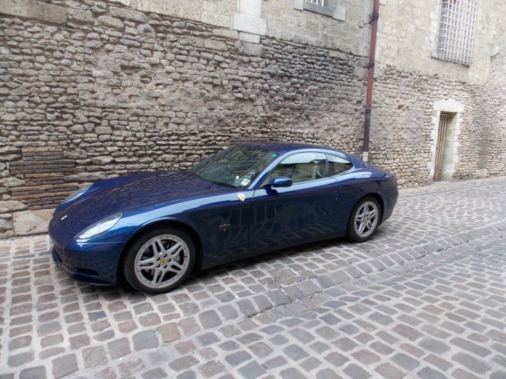 Manual 612 at RM auctions - Page 1 - Ferrari V12 - PistonHeads - The image features an **indigo blue** luxury sports car parked by a **rock-walled building**. The car is a **Ferrari F12**, a high-performance vehicle known for its sleek design and powerful engine. The setting appears to be a **narrow street**, with the building and the road providing a picturesque backdrop. The stone riverbed further enriches the overall image, creating a sense of tranquility amid the luxury. The car's position at the corner of the street gives the impression that it's ready to speed off into a distance.