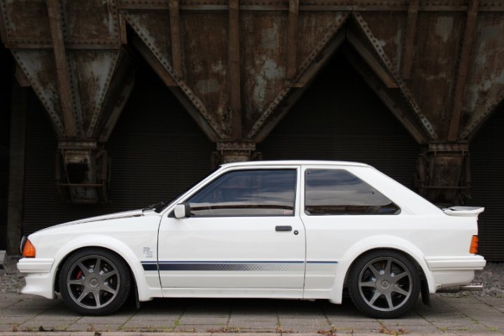 Longest ownership and still yours - Page 4 - Classic Cars and Yesterday's Heroes - PistonHeads - The image shows a white sports car parked indoors, possibly in a warehouse or a similar setting. It features a modified rear spoiler and side skirts, with the body showing various aerodynamic additions. There's also a black roof rack. The vehicle has tinted windows and is adorned with aftermarket accessories such as decals, badges, and possibly stickers. The car appears to be in good condition, suggesting it may be well-maintained or restored. The surroundings indicate an urban setting due to the industrial backdrop and the concrete flooring.