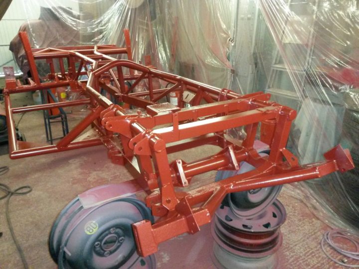 A body off in pictures, LOTS of pictures. - Page 1 - S Series - PistonHeads - The image depicts a mechanical structure that appears to be under construction or restoration. A red chassis is prominently visible, featuring two large, hollow sports car-like rear panels and a matching set of smaller front panels. The chassis sits in a room with plastic sheeting and adhesive tape that indicates it's being prepared for something, possibly painting or reshaping. In the background, there are metal racks and what looks like a floor plan or design blueprint, suggesting a manufacturing or customization setting.