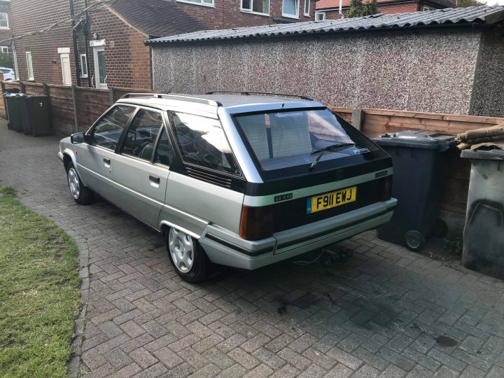 Citroen BX 1.9D 'The Estate' - Barn Stored to Bahn Stormer? - Page 16 - Readers' Cars - PistonHeads - The image is a photograph displaying a gray and black compact car parked beside a bricked driveway. The vehicle has a European license plate with the text "F51 BPW". The front left wheel is slightly askew, indicating possible damage or prior movement.

In the background, there is access to a residential area with a paved path leading to a house. On the right side of the image, a black trash bin is visible beside the car. There is also a glimpse of a metal chicken wire fence in the right background, partially hidden by the trash bin.

The lighting and shadows in the image suggest it is taken outdoors during daytime under overcast or diffuse lighting conditions. There are no visible texts or distinctive markings aside from the license plate of the car, which is in focus.