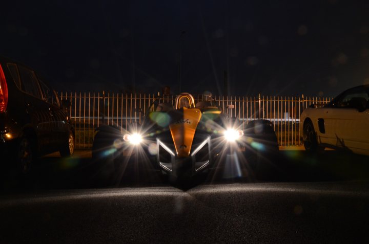 Essex Pistonheads March - The image captures a dramatic night scene with a large vehicle parked in front of its container. The vehicle is a prominent feature, with its yellowish hue and bright headlights shining on the surroundings. The sky is dark, and the tracks in the foreground lead towards a metal gate, adding a sense of depth and mystery to the scene.