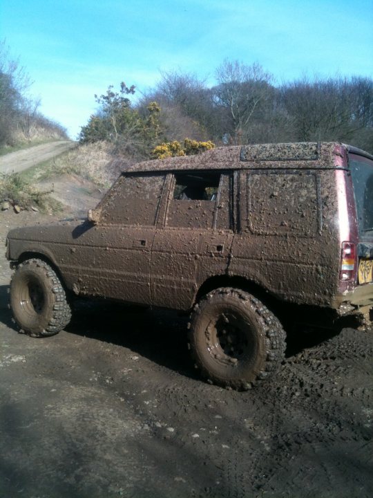 Wilton off road centre pics 11/03/2012 - Page 1 - Off Road - PistonHeads - The image shows a brown, mud-covered vehicle, likely a four-wheel-drive SUV, possibly parked on the side of a road. The wheels are buried in deep, wet mud, indicating that it has been used off-road or during a period of heavy rain. The roof and hood are also coated with mud, further suggesting off-road use. The surrounding environment looks like a natural landscape with green bushes and patches of trees, but it is mostly obscured by the mud on the vehicle. There seems to be a dirt road or a path visible beside the vehicle. The car appears to be out of use, as the mud has entirely covered it, making it difficult to discern any details about the vehicle beyond its color and the presence of roof racks.