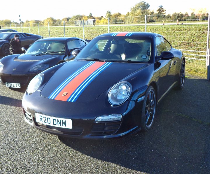 A car parked on the side of the road - Pistonheads - In the image, a black Porsche car is parked on an asphalt surface, positioned diagonally from the viewer's perspective. The car features a red and blue stripe that runs along the hood, partially covering a small part of the front grille. The registration plate is clearly visible with the letters "R20 DNM" printed on it. In the background, other cars are visible, suggesting the image might have been taken in a public parking area. The scene gives off a serene and peaceful atmosphere, with a field visible in the distance under a clear sky.