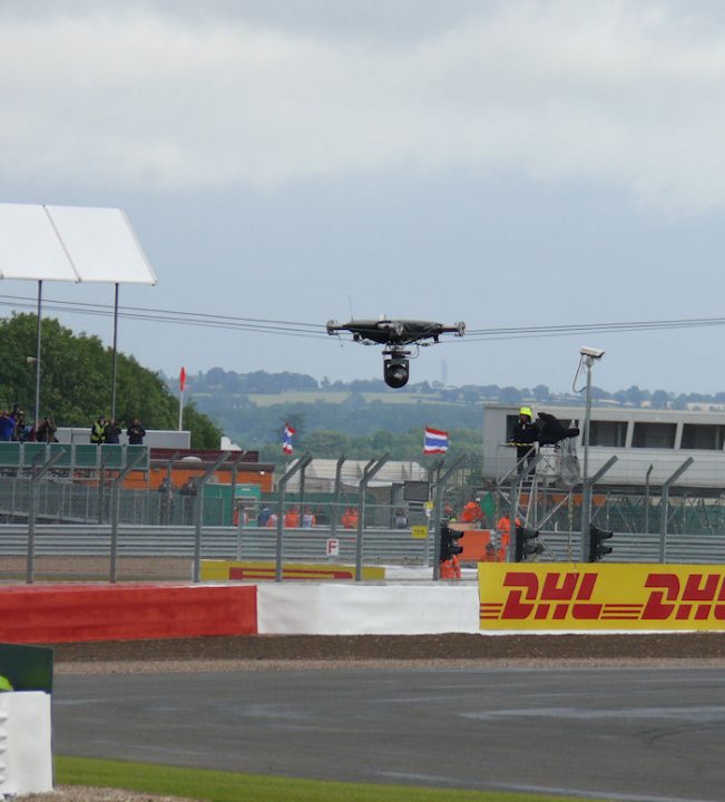 The Official British Formula One GP ***Spoilers*** - Page 96 - General Motorsport - PistonHeads - The image captures an exciting moment at a motorsport event, featuring a black racing drone hovering above a racetrack. In the foreground, a driver can be seen, gearing up for a lap. The track itself is a vibrant mix of orange, white, and several other stripes, with red and white barriers lining its edges. The background is adorned with lush green hills and trees, adding a touch of nature to the thrilling setting of the race. A crowd of spectators, possibly at a stand or in their viewing spots, are watching the spectacle.