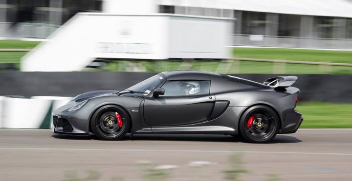 A black car parked next to a parking meter - Pistonheads - The image is a dynamic, motion-blurred photograph of a sports car in action on a track. The car is predominantly dark in color, specifically black and grey, suggesting a high-performance model likely designed for speed and aerodynamics. It features aggressive styling with clean, angular lines, wide arches, and various aerodynamic components such as a spoiler, trunk spoiler, and a side splitter. The car is captured in mid-corner, with the front-end out of view, implying a high level of speed. The background is blurred but appears to be a racetrack with a grassy area, a building, and a wall, which helps to emphasize the motion of the car. The wheels are black with red accents, adding to the car's aggressive look.