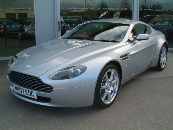 Hello All - Page 1 - Aston Martin - PistonHeads - The image showcases a silver Aston Martin sports car. It's a modern vehicle with the characteristic design of Aston Martin, known for its sleek lines and aerodynamic prowess. The car is parked in a lot with other cars in the background, suggesting it's being displayed or possibly sold. The car's registration is visible, reading "NK57 EDC". The license plate is clearly displayed for all to see.