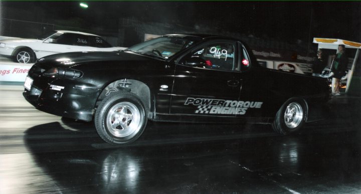 Prototype Driveshaft - Page 2 - HSV & Monaro - PistonHeads - The image shows a black vehicle with text that reads "POWERtorque" on the side, suggestive of its customized nature labelled by a racing team. It's captured in action, driving on a surface where the car casts a long, dark shadow and there's visible motion blur. The vehicle is racing or test driving in a sportier setting, as indicated by the presence of other vehicles and lights in the background, implying a race track or darked event.