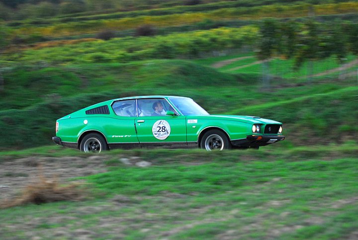 Americanaustralian Pistonheads - The image captures a vibrant scene of a green vintage car racing along a verdant hillside. The car, exposed to the elements, is racing with its hood up, adding a sense of motion and excitement. The hillside itself is lush and green, dotted with trees that frame the car's journey. In the distance, fields sprawl under the open sky, further enhancing the sense of speed and adventure. The car is driving towards the left side of the image, leaving a trail of green in its wake.