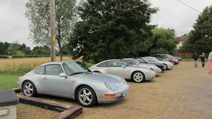 Hare Breakfast Meet - August - Page 1 - Kent & Essex - PistonHeads - The image displays a parking lot with a variety of cars parked in a line. In the foreground on the left, there is a silver-colored Porsche 944 with a distinctive black side stripe. Next to it, other cars are visible, but the specific model, make, or color of these cars is not clearly discernible. The setting seems to be outdoors with partial views of trees and what looks like a grassy area beyond the parking lot. There are no visible people or personal objects like handbags in the immediate vicinity of the cars. The photo is taken at an angle that provides a clear view of the front cars in the lineup, but the further back cars are only partially visible. The lighting suggests it is a cloudy day.