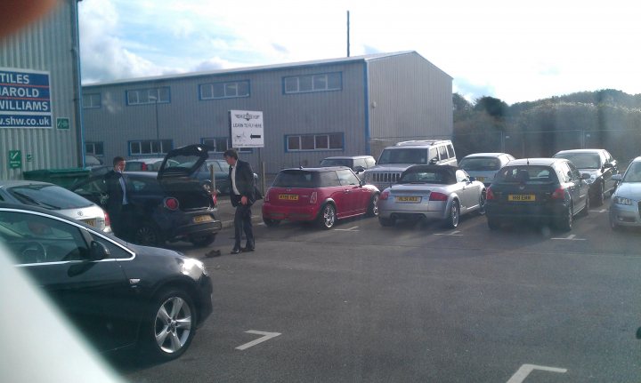 The BAD PARKING Thread [Vol 2] - Page 149 - General Gassing - PistonHeads - The image captures a scene in a parking lot in the UK, where a variety of cars are parked. There's a red car in the center of the frame, and a black car to its left. A blue car is visible to the right of this black car. The parking lot is lined with a long building, and there's a white sign standing near the cars as if attracting attention. It appears to be a clear day with plenty of light illuminating the scene.